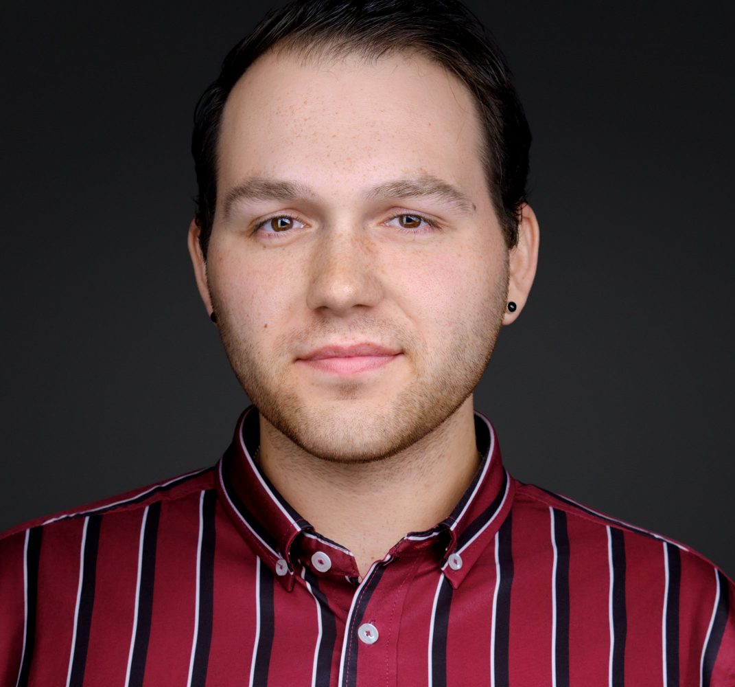 Man with short hair in a red striped shirt against a dark background.