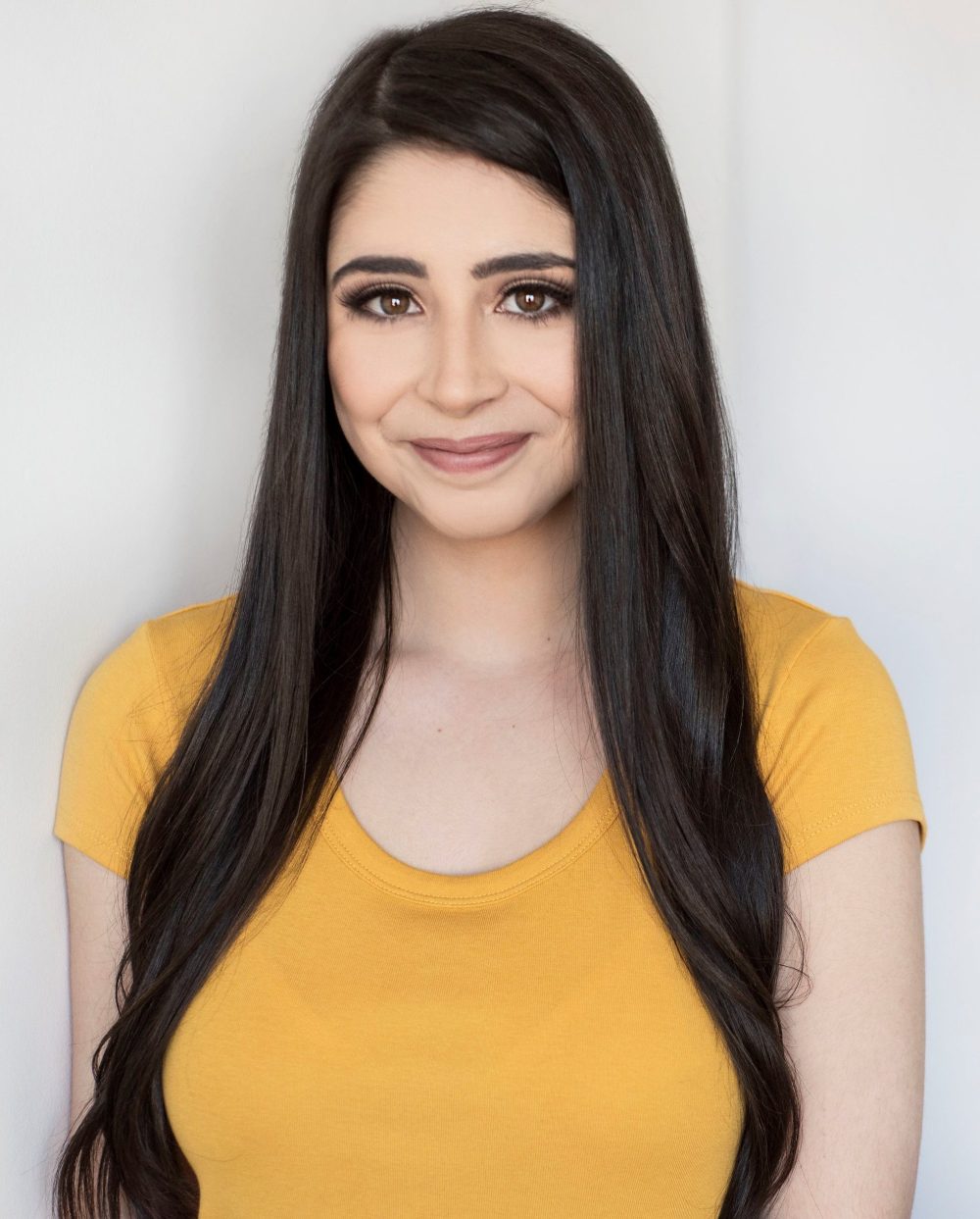 Woman with long hair in a yellow shirt smiling against a plain background.