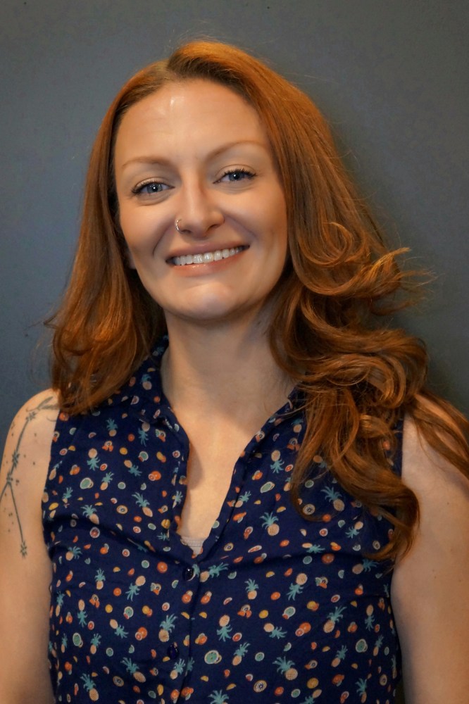 Smiling woman with long red hair and a patterned blouse against a plain background.
