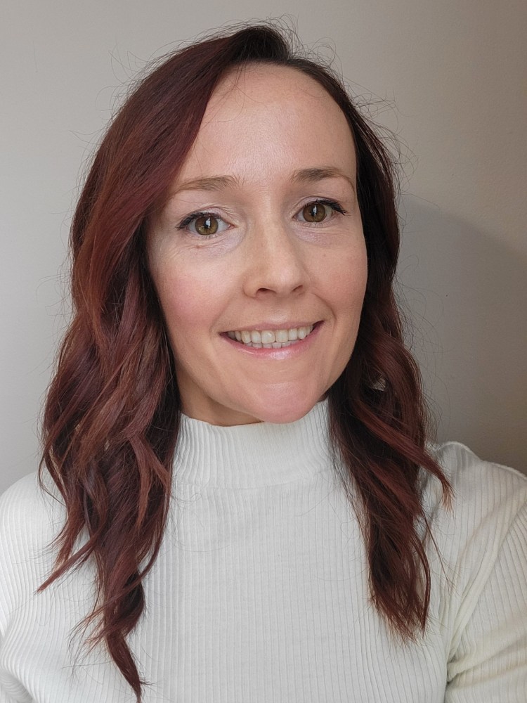 Woman with wavy brown hair smiling, wearing a white turtleneck.