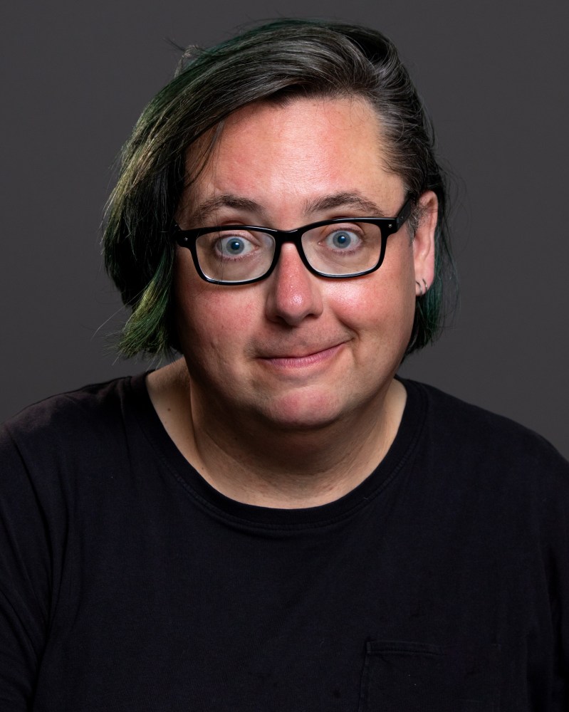 Person with glasses and short hair smiling, wearing a black shirt against a dark background.