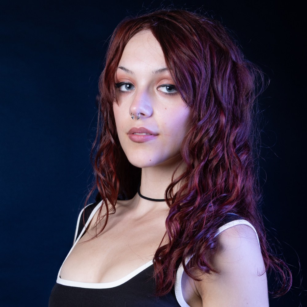 Person with long, wavy red hair, wearing a black and white top against a dark background.