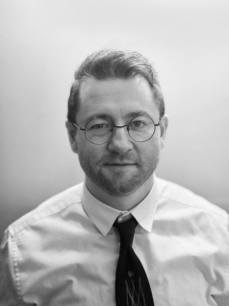 Man in glasses, white shirt, and tie, with a neutral expression, in grayscale photo.
