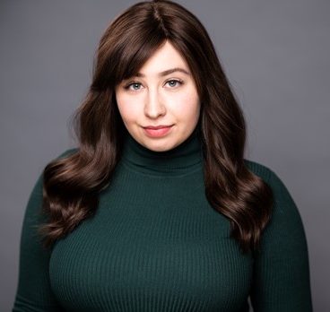 Woman with long brown hair in green turtleneck against gray background.